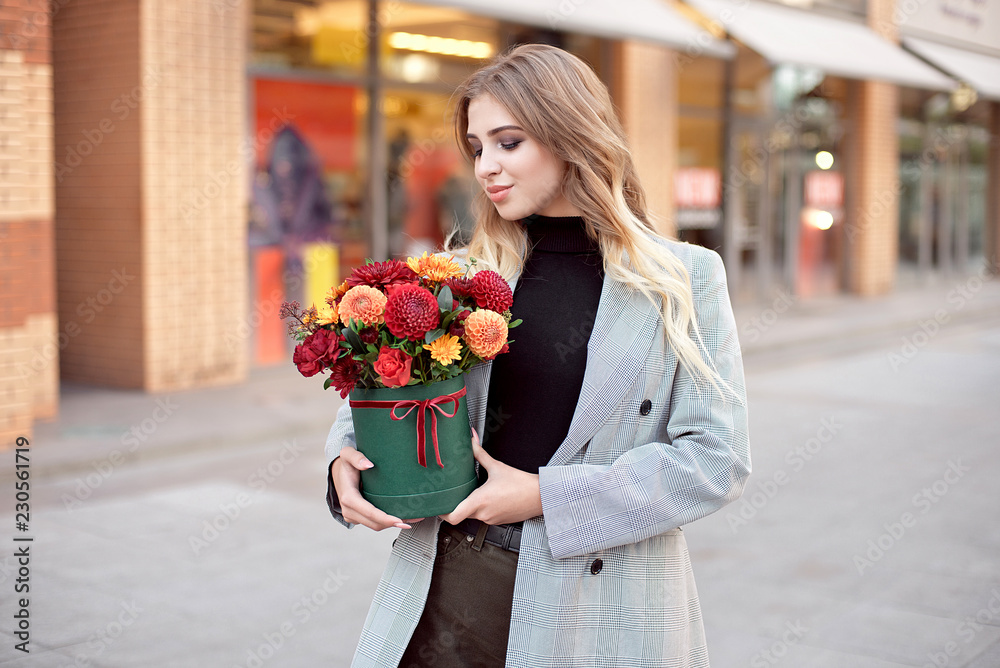 Fototapeta premium Caucasian fashion woman standing on the street near storefront shop windows holding flower-box with happy smile.