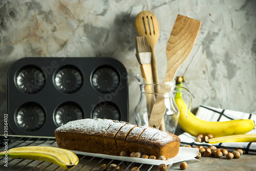 Sliced banana bread is lying on a board among kitchen utensils, bananas and hazelnuts. Homemade bakery concept photo