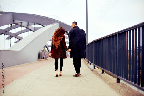 Young romantic couple, beautiful girl with flowers has dating in the city. Man in gray overcoat and model in red coat. Loving happy, smile, enjoy a walk together on a autumn day on the bridge of lover