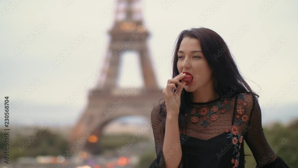 Paris woman smiling eating the french pastry macaron in Paris. Evening Eiffel tower with lights in the background. Portrait of gorgeous romantic young sensual girl in fashion black sexy dress enjoying
