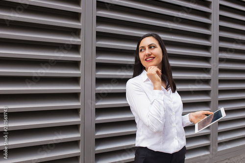 Portrait of flirty businesswoman with pc tablet looking away. Young Caucasian female student standing with digital tablet outdoors. Businesswoman concept