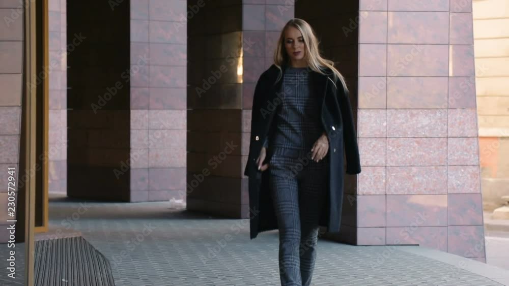 Successful woman in gray casual outfit and black smart coat walking along the lane, outdoor shoot near large morden building Successful woman in gray casual outfit and black smart coat walking along the lane, outdoor shoot near large morden building