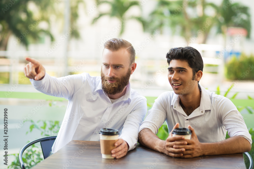 Two male friends drinking coffee in outdoor cafe. One man showing something away with blurred plants in background. Coffee break concept. Front view. Two male friends drinking coffee in outdoor cafe. One man showing something away with blurred plants in background. Coffee break concept. Front view.