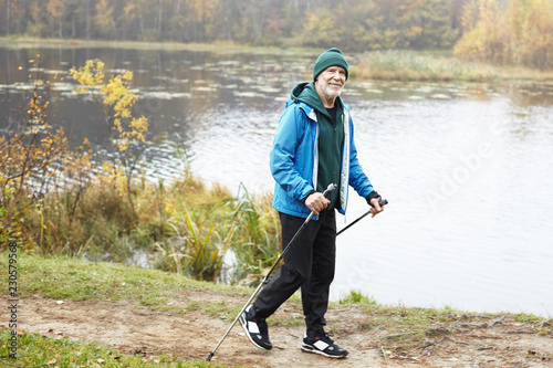 Full length portrait of elderly bearded male trekker enjoying nordic walking outdoors, using poles, passing by pond in background. Senior seventy year old man training outside in wild nature