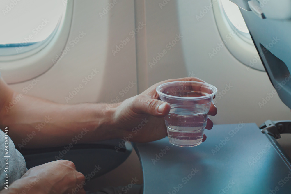 Fototapeta premium Passenger drinking water in airplane during flight. Close up of hand holding glass in plane.