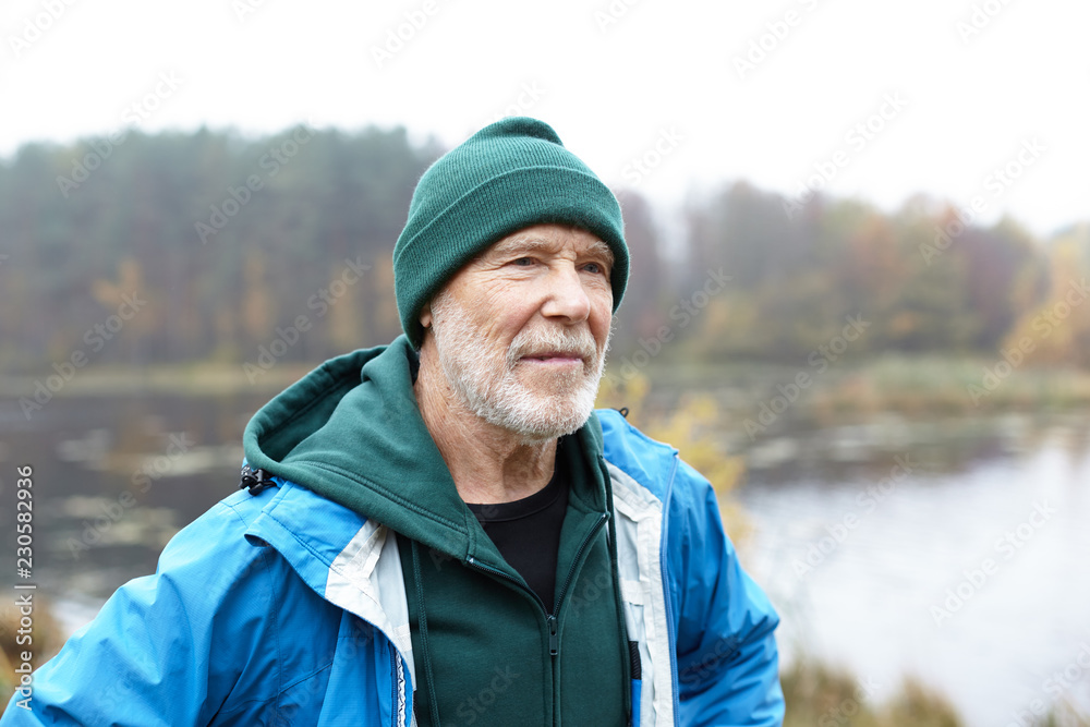 Portrait of senior seventy year old man with gray stubble and wrinkled face thinking about something outdoors, standing by river while walking alone on autumn day, wearing hat and jacket over hoodie