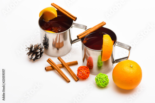 Mugs with mulled wine or hot drink near orange fruit on white background, close up. Mulled wine or hot beverage in metal mugs with cinnamon sticks and fir cone. Winter hot drinks concept