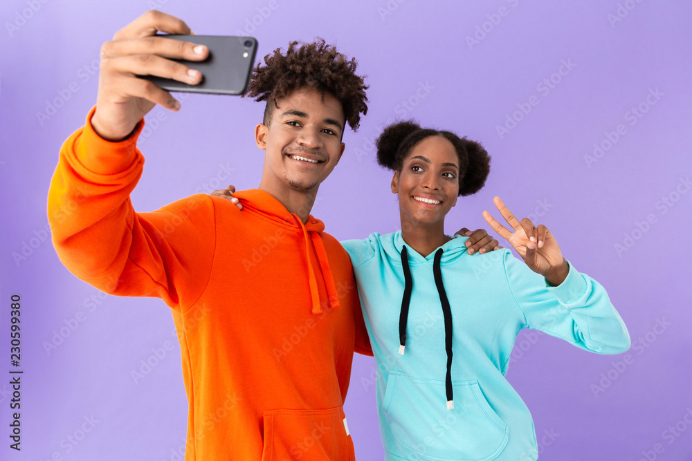 Photo of happy african american couple wearing colorful sweatshirts taking selfie on smartphone, isolated over violet background Photo of happy african american couple wearing colorful sweatshirts taking selfie on smartphone, isolated over violet background