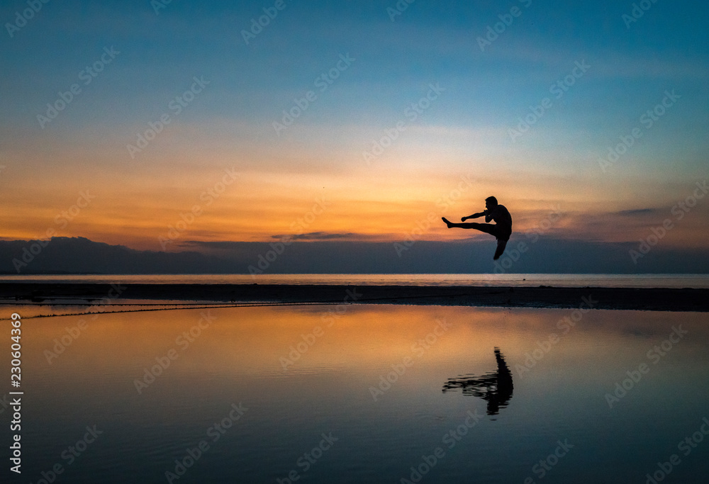 young athletic guy performing a flying kick during sunset on the beach