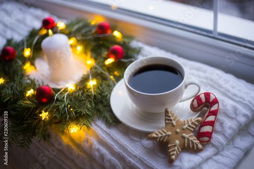 White cup of coffee and Christmas gingerbread near fir wreath decorated with red balls, burning candle and coiled with glowing garland with warm light near window.