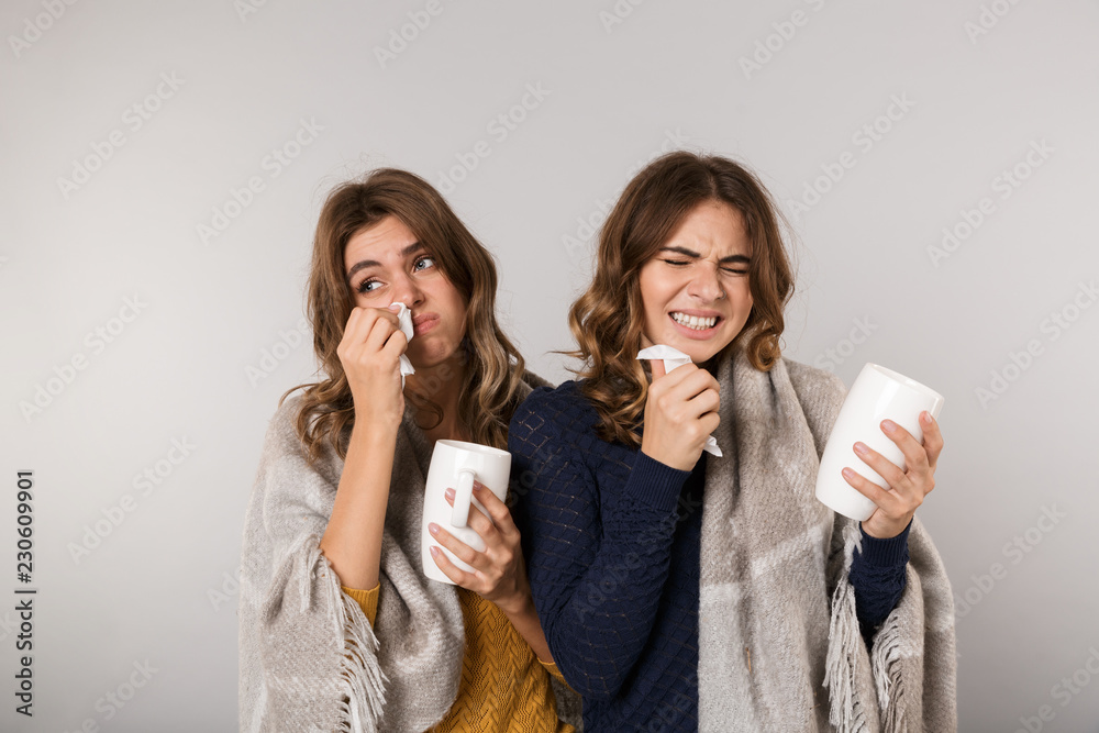 Image of two ill women covered with blanket drinking hot tea from cups, isolated over gray background
