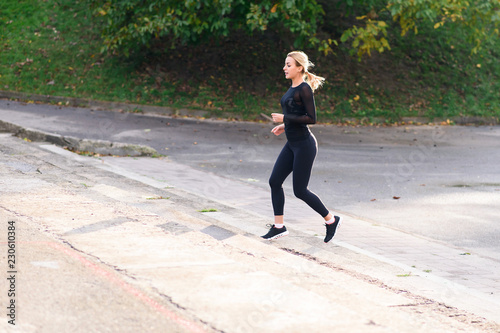 Young attractive fit woman in black sportswear jogging the stairs in the park. Blonde girl training and working out outdoors.