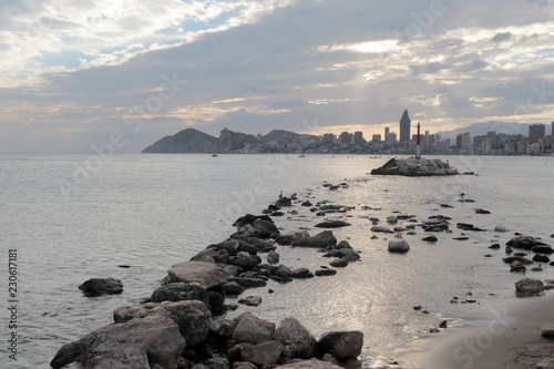 Meditarranean sea landscape in Benidorm at sunset. Rocks and the lighthouse on a small island with buildings in the background on a cloudy day.
