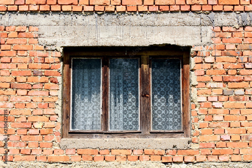 Old dilapidated house window with white curtain and cracked faded wooden frame on red brick wall on warm sunny day