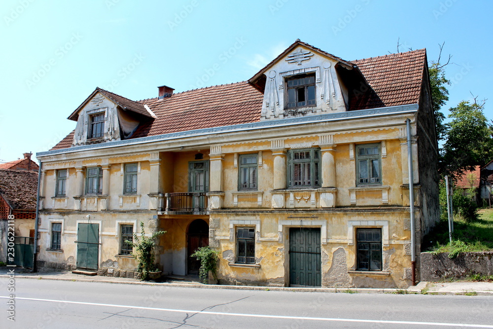 Old large abandoned baroque style family house with cracked dilapidated facade, broken windows and locked doors next to asphalt road with clear blue sky in background