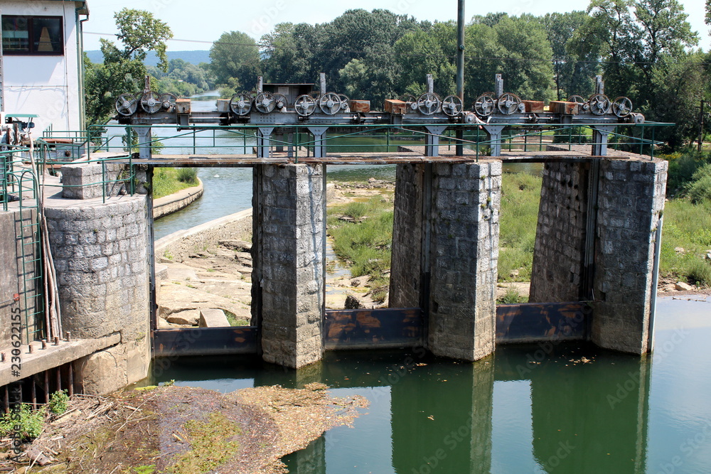 Old small dam made of stone and metal used to regulate and direct water to hydropower plant building surrounded with low water level with dry grass in front and river with tall trees in background on Old small dam made of stone and metal used to regulate and direct water to hydropower plant building surrounded with low water level with dry grass in front and river with tall trees in background on