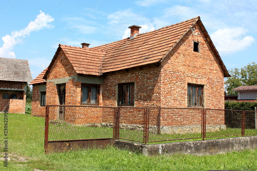 Unfinished red brick family house with worn down doors and faded window frames surrounded with high uncut grass and rusted metal fence with hedge and cloudy sky in background Unfinished red brick family house with worn down doors and faded window frames surrounded with high uncut grass and rusted metal fence with hedge and cloudy sky in background