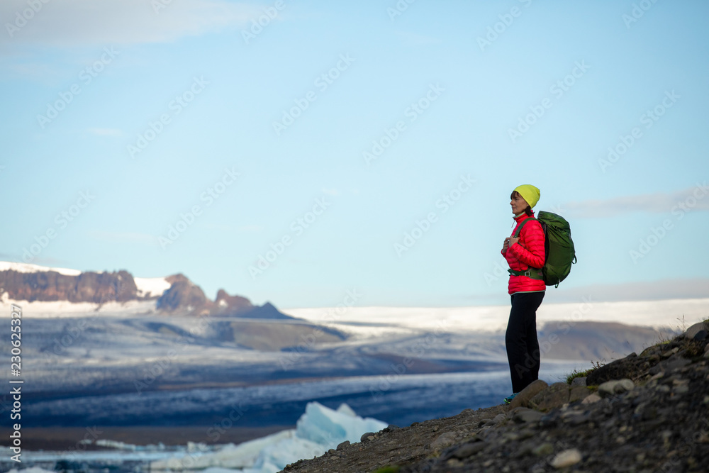 Young sporty girl with backpack admires beautiful view of the ice lake and mountains. Ice lagoon in Iceland