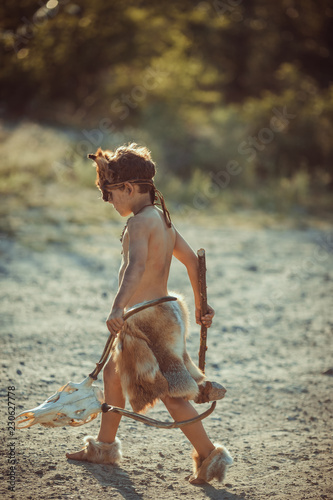 Angry caveman, manly boy with stone axe and animal skull. Prehistoric tribal boy outdoors on nature. Young shaggy and dirty savage, warrior and hunter with weapon. Primitive ice age man in animal skin photo