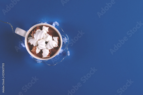 Hot chocolate with marshmallow candies decorated illumination light on blue paper background. Top view. Copy space. Toned