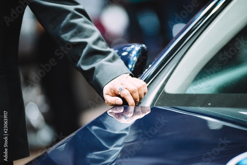 An unidentified man in a black vest holding a notebook was walking across a black modern car. Handsome elegant man in suit, businessman or salesman, stretches his hand to conclude a business.