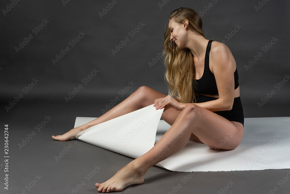 Obraz premium A beautiful smiling young pregnant woman blonde wearing a black maternity underwear on a gray background sits on a large sheet of white paper and tears it. Fashion, health, beauty.
