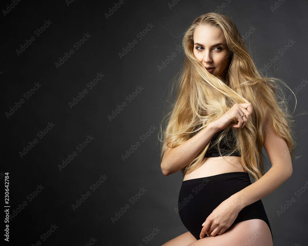 Beautiful pregnant leggy blonde woman wearing black maternity underwear sits on a chair on a gray background and touches her hair. Health, fashion, motherhood. Commercial design. Beautiful pregnant leggy blonde woman wearing black maternity underwear sits on a chair on a gray background and touches her hair. Health, fashion, motherhood. Commercial design.