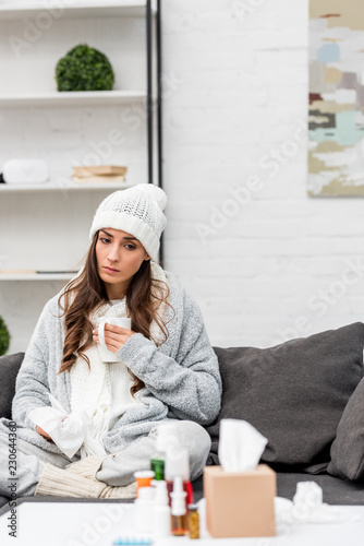 sick young woman in warm clothes at home holding cup of tea and looking at various medicines standing on table on foreground
