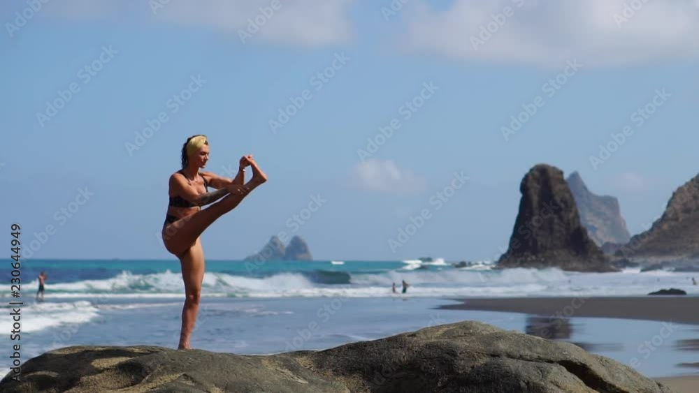 Young girl in bikini balancing standing on one leg doing yoga standing on a rock by the ocean on a black sand beach Young girl in bikini balancing standing on one leg doing yoga standing on a rock by the ocean on a black sand beach