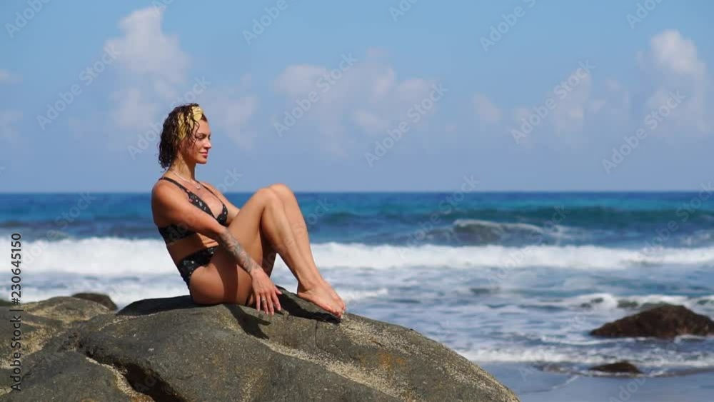 A young girl in a bikini balancing sitting on a stone and doing exercises for the abdominal muscles alternately lifting her legs. Training on the background of the ocean on the black sand beach A young girl in a bikini balancing sitting on a stone and doing exercises for the abdominal muscles alternately lifting her legs. Training on the background of the ocean on the black sand beach