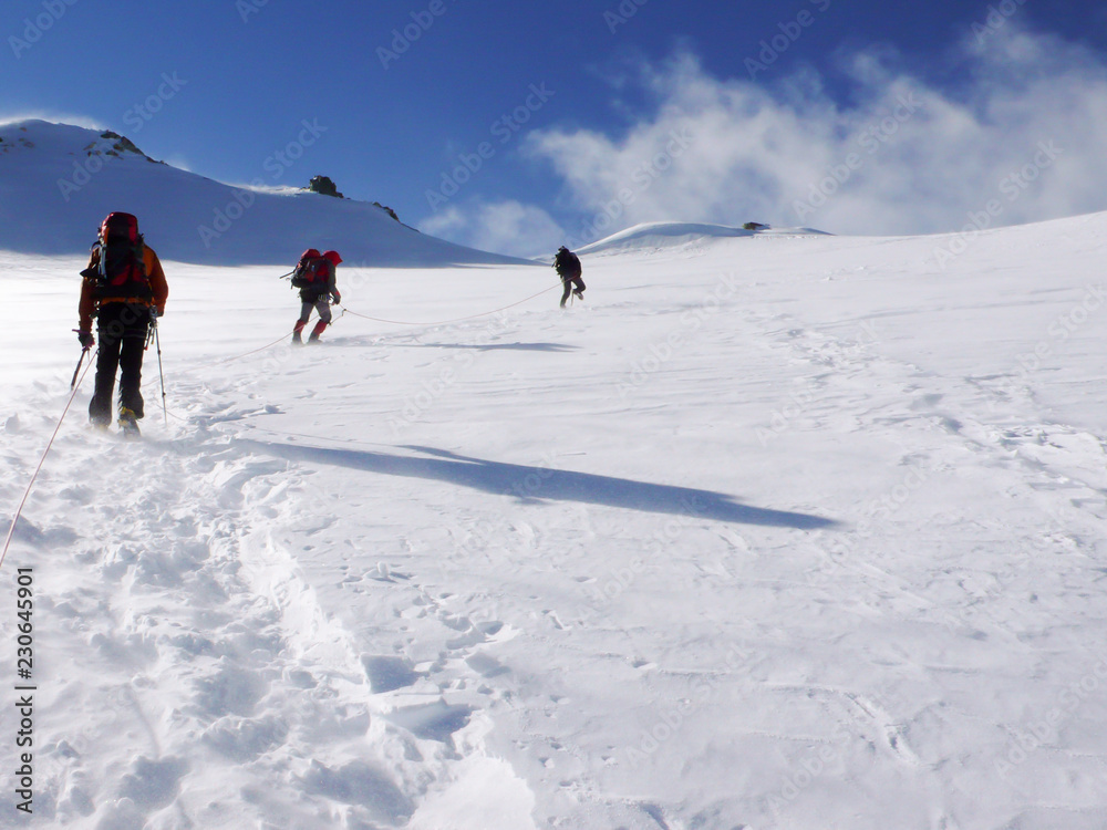 three mountain climbers cross a high alpine glacier in windy stormy weather on their way to a mountain peak far ahead
