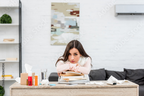 sick young businesswoman leaning on stack of books at workplace and looking at camera