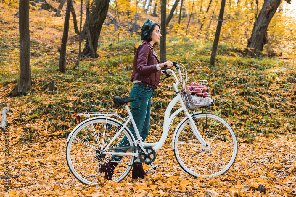 side view of young woman in stylish leather jacket and beret carrying bicycle in yellow forest