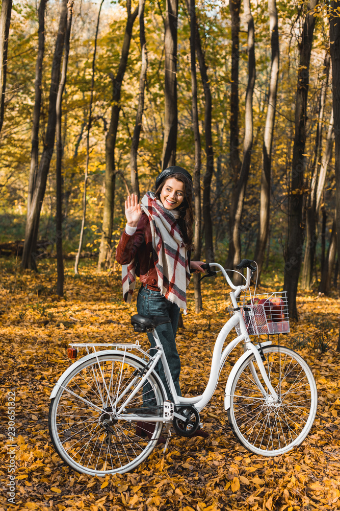 smiling stylish girl in leather jacket and beret waving by hand and carrying bicycle in autumnal park smiling stylish girl in leather jacket and beret waving by hand and carrying bicycle in autumnal park