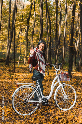 smiling stylish girl in leather jacket and beret waving by hand and carrying bicycle in autumnal park