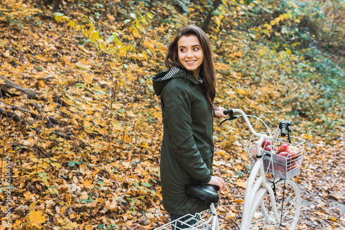 smiling young woman carrying bicycle with basket full of apples in yellow autumnal forest