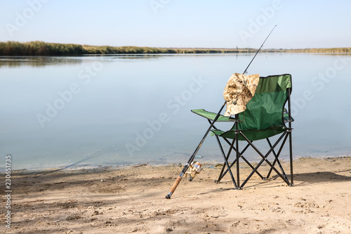 Camping chair and fishing rod at riverside on sunny day. Space for text