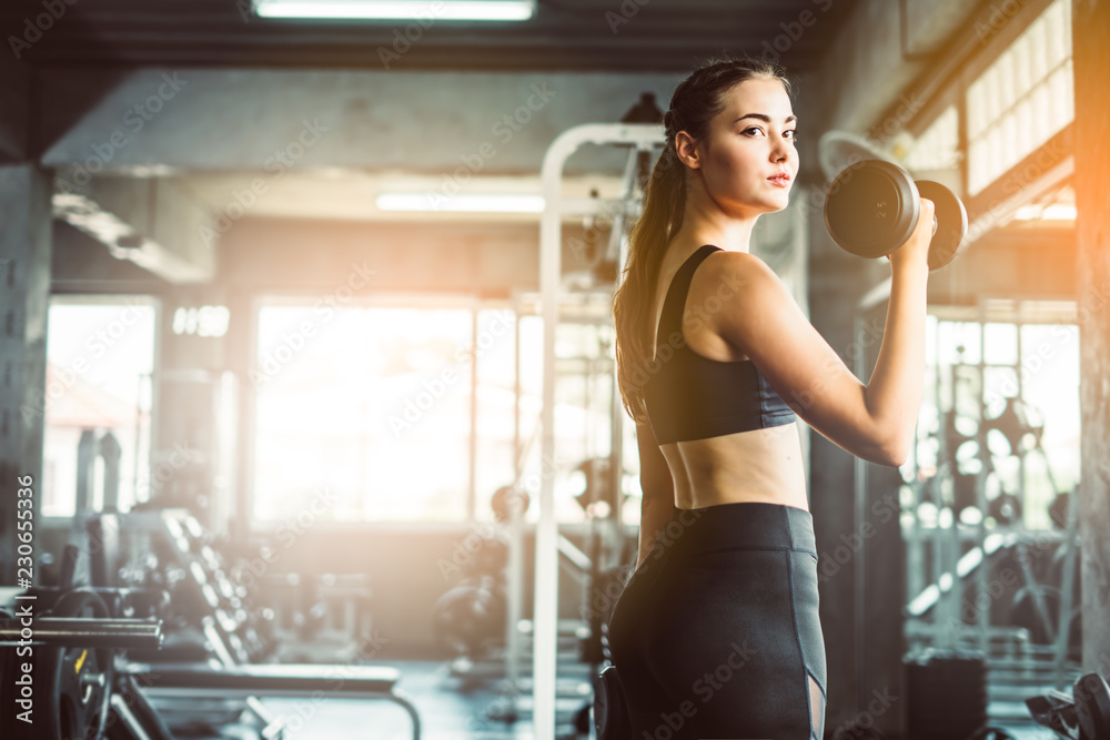 Fototapeta premium Young girl playing dumbbell to exercise in fitness.Slim girl lifts heavy dumbbell while training in the gym. Sports concept fat burning and a healthy lifestyle.