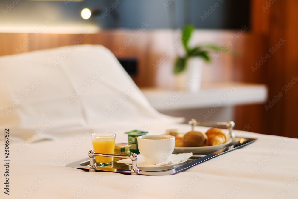 Plateau de petit-déjeuner contenant du café, jus d'orange, croissant et  yaourt posé sur le lit d'une chambre d'hôtel recouvert d'un drap blanc avec  une plante verte en arrière plan Stock Photo