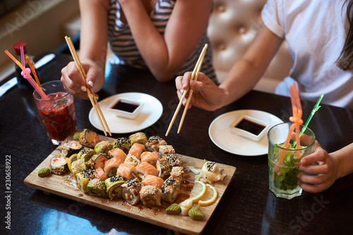 Two yang girls are eating delicious rolling sushi at restaurant served on the wooden board with chopsticks, soy sauce and cocktails