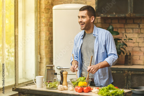 It's so delicious! Casual happy young man preparing salad at home in loft kitchen and smiling.