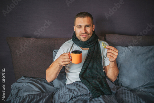 Sick but positive young man sits on bed in bedroom and looks on camer. He holds orange cup in one hand and piece of lemon in another one. Guy smiles. He looks happy. photo