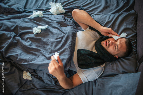Picture of young man lying on bed and suffering from headache and nose running. He feels bad and exhausted. Young man holds dry napkin on head and shrinking. There are lots of napkins on bed.