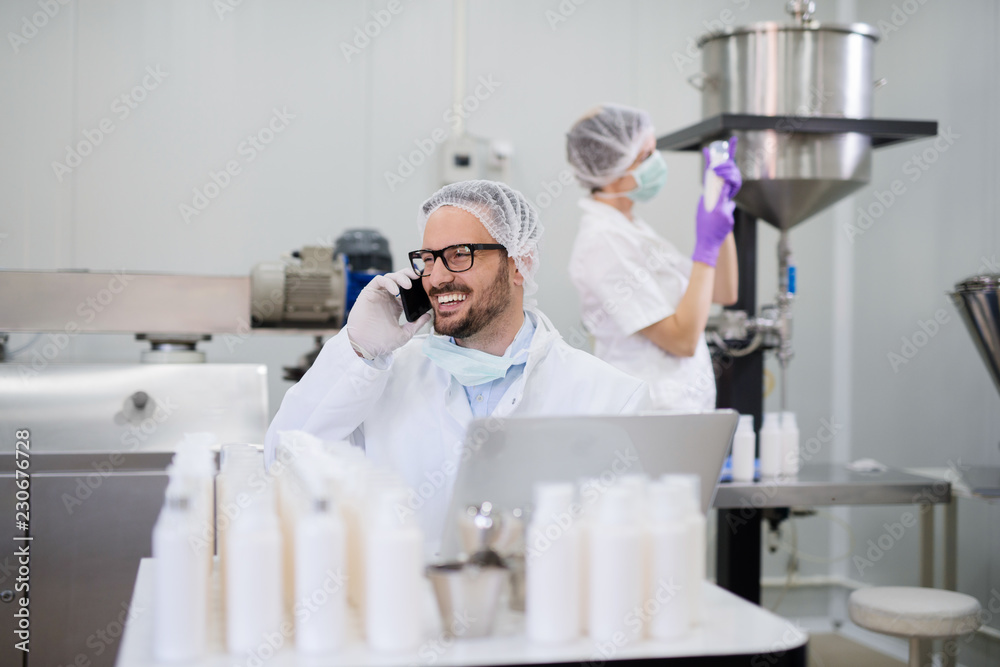 Man in working suit talking on the mobile while sitting in the lab. In background his assistant holding cosmetic product. Man in working suit talking on the mobile while sitting in the lab. In background his assistant holding cosmetic product.