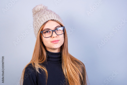 Young beautiful girl with glasses and winter hat standing in front of grey background, a lot of clean space