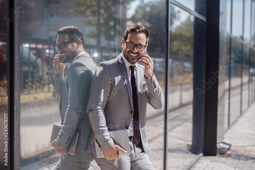 Portrait of smiling businessman using smart phone while leaning on window. In other hand tablet.