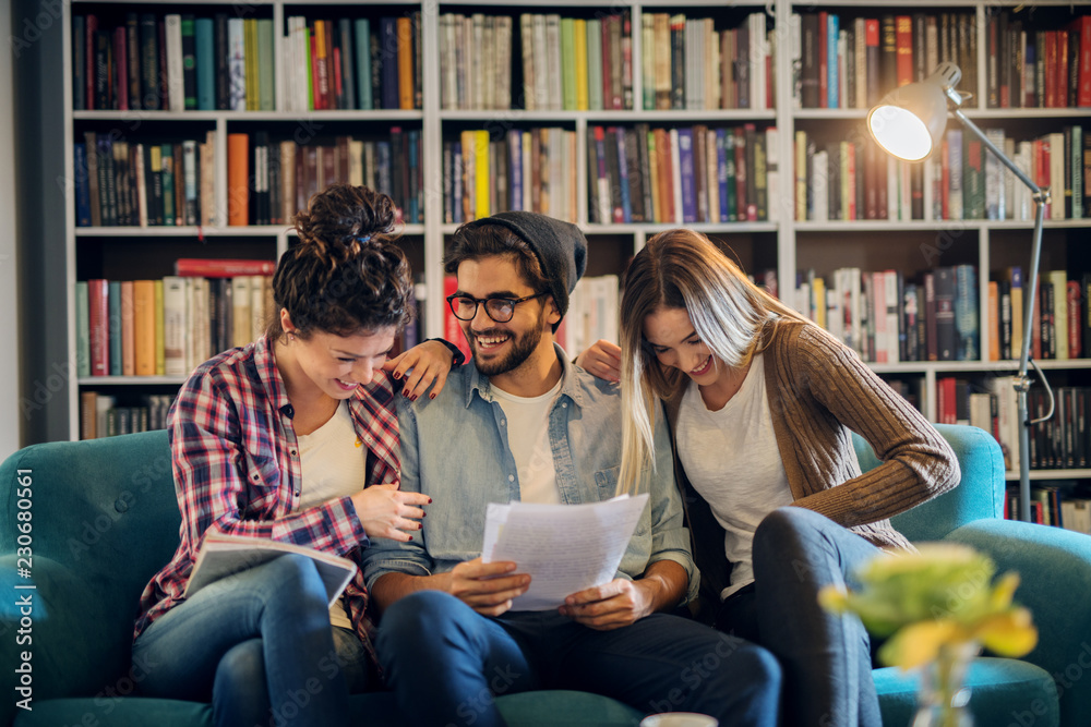 Group of three young students sitting in library and preparing exam together. Cheerful teenagers having fun and learning. Group of three young students sitting in library and preparing exam together. Cheerful teenagers having fun and learning.