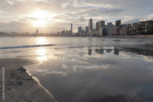 Landscape on the shore of the mediterranean sea in Benidorm on the Poniente beach with buildings, the sun at sunset and the clouds reflected in the water