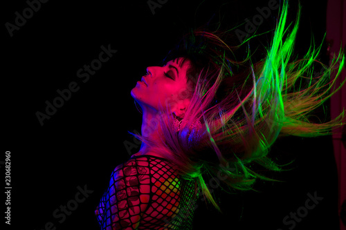 Alternative model with bangs and colored hair poses under pink, blue, and green light wearing a fishnet top