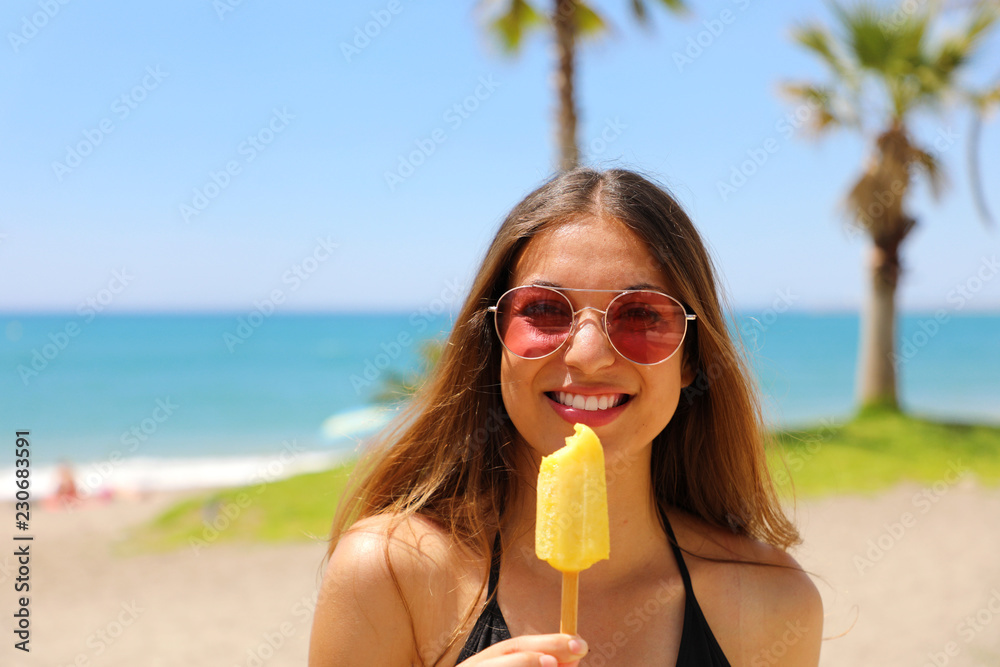 Smiling beautiful girl with sunglasses on the beach eating ice lolly with palm trees on the backgroud. Summer holidays concept. Smiling beautiful girl with sunglasses on the beach eating ice lolly with palm trees on the backgroud. Summer holidays concept.